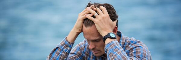 A man in a plaid shirt sits by the water looking distressed, symbolizing stress.